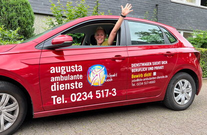 Mitarbeiterin winkt aus rotem Fahrzeug der Ambulanten Pflege