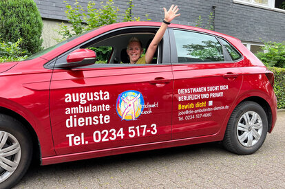 Mitarbeiterin winkt aus rotem Fahrzeug der Ambulanten Pflege